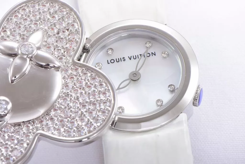 Louis Vuitton Women's Watch Vivienne Bijou Secret, Vivienne Mascot Collection Movement: Quartz. Functions: hour and minute display. Case: 18-carat white gold, 18-carat pink gold, 18-carat yellow gold with diamonds. Diameter: 34 mm. Anti-reflective sapphire crystal. Water-resistant to 30 m. For the first time, LOUIS VUITTON has incorporated its mascot, Vivienne, into the world of watchmaking, with the brave and charming Vivienne doll, dressed in diamond-embellished gold, to create a unique ladies' watch: the Vivienne Bijou Secret. Vivienne's face is movable, covering or revealing the 21 mm white gold case between rotations. The dial, appliquéd with white mother-of-pearl, is adorned with a graceful signature Monogram motif and set with diamond hour markers. The watch is currently available on four straps in red, black, green and white leather.